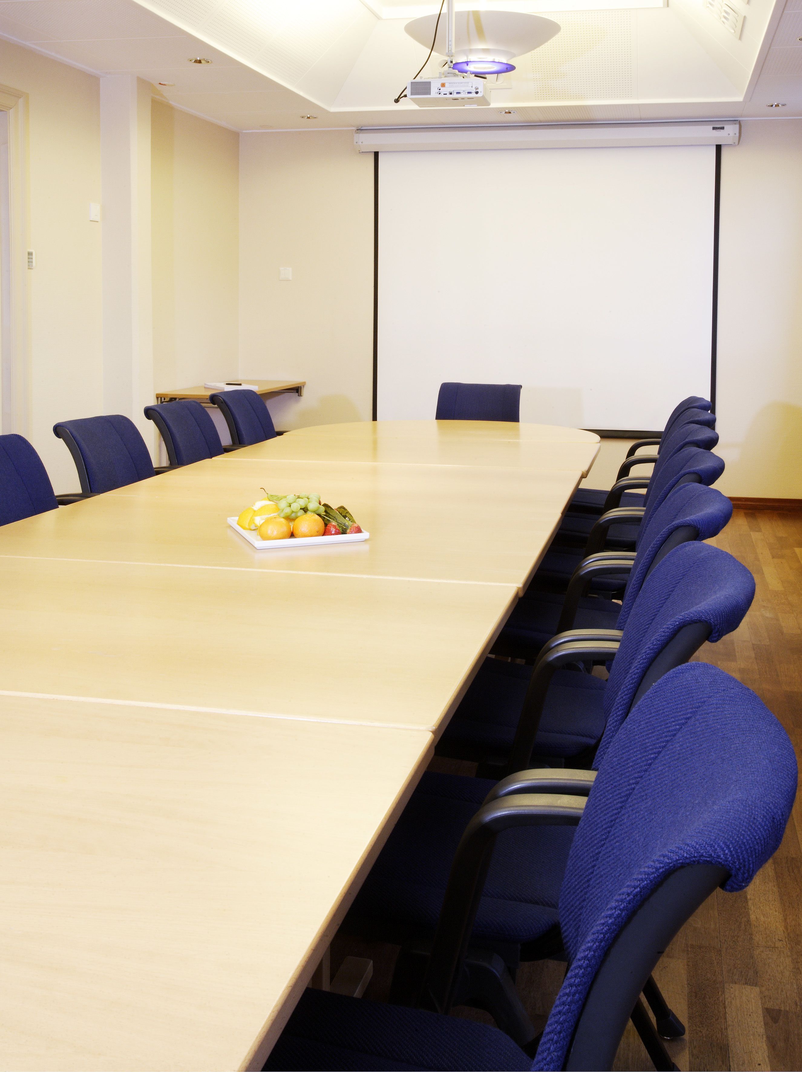 Een vergaderzaal met een lange tafel, blauwe stoelen en een projectiescherm.