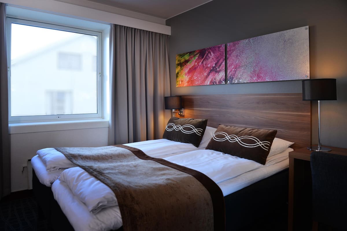 A hotel room with a large bed, a window with curtains, a wall-mounted artwork, and a bedside table with a lamp.