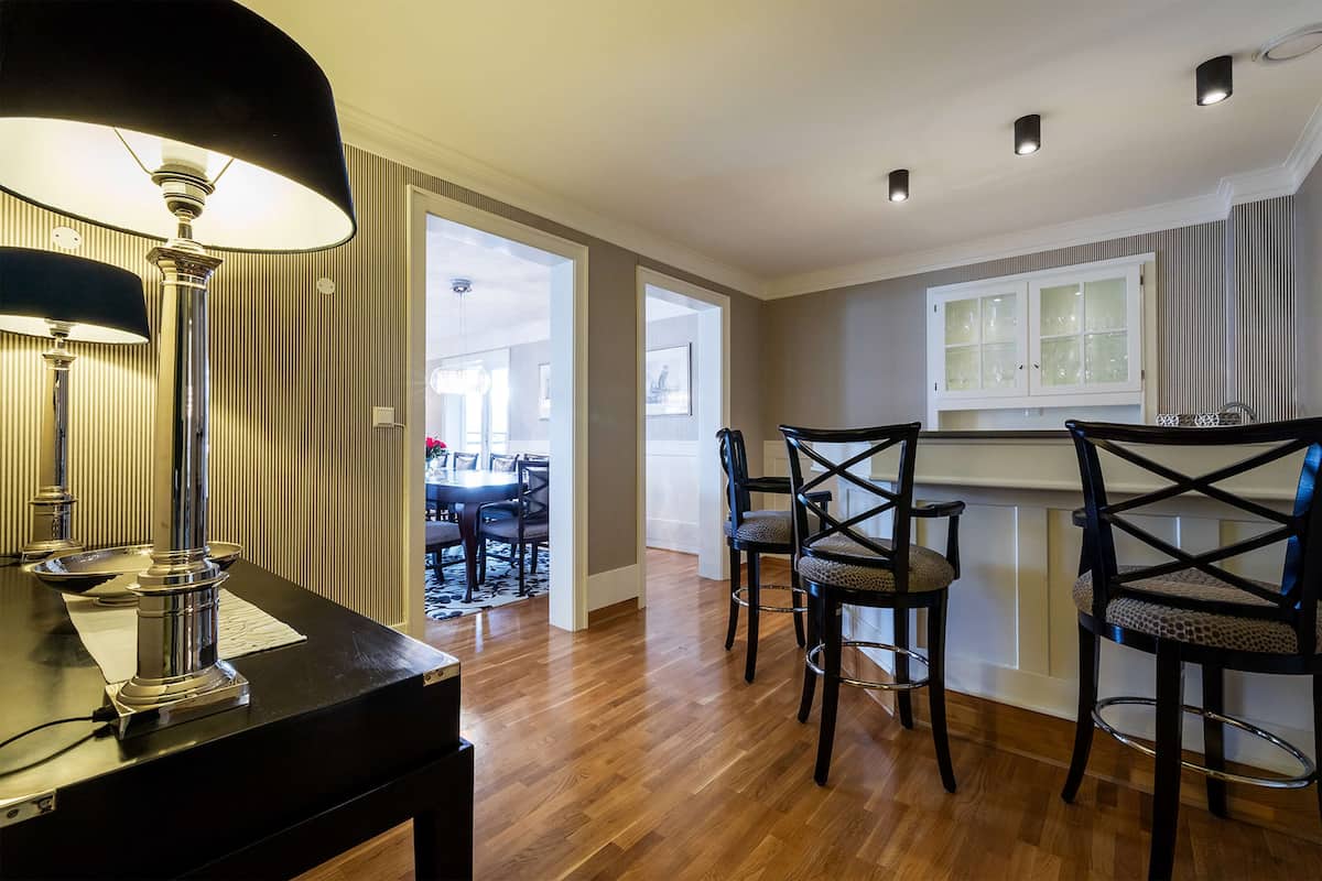 A dining area with a black table, two chairs, and a lamp. There is a window with curtains and a door leading to another room.