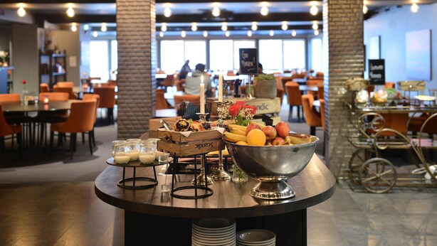 A buffet area with a variety of food items, including fruits and pastries, in a modern restaurant setting with brick walls and orange chairs. - Scandic Fauske Hotel (Fauske)