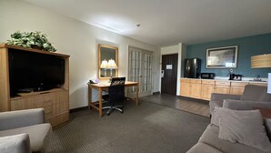Habitació d'hotel amb televisió, escriptori, cadira, sofà, zona de cuina i una porta.