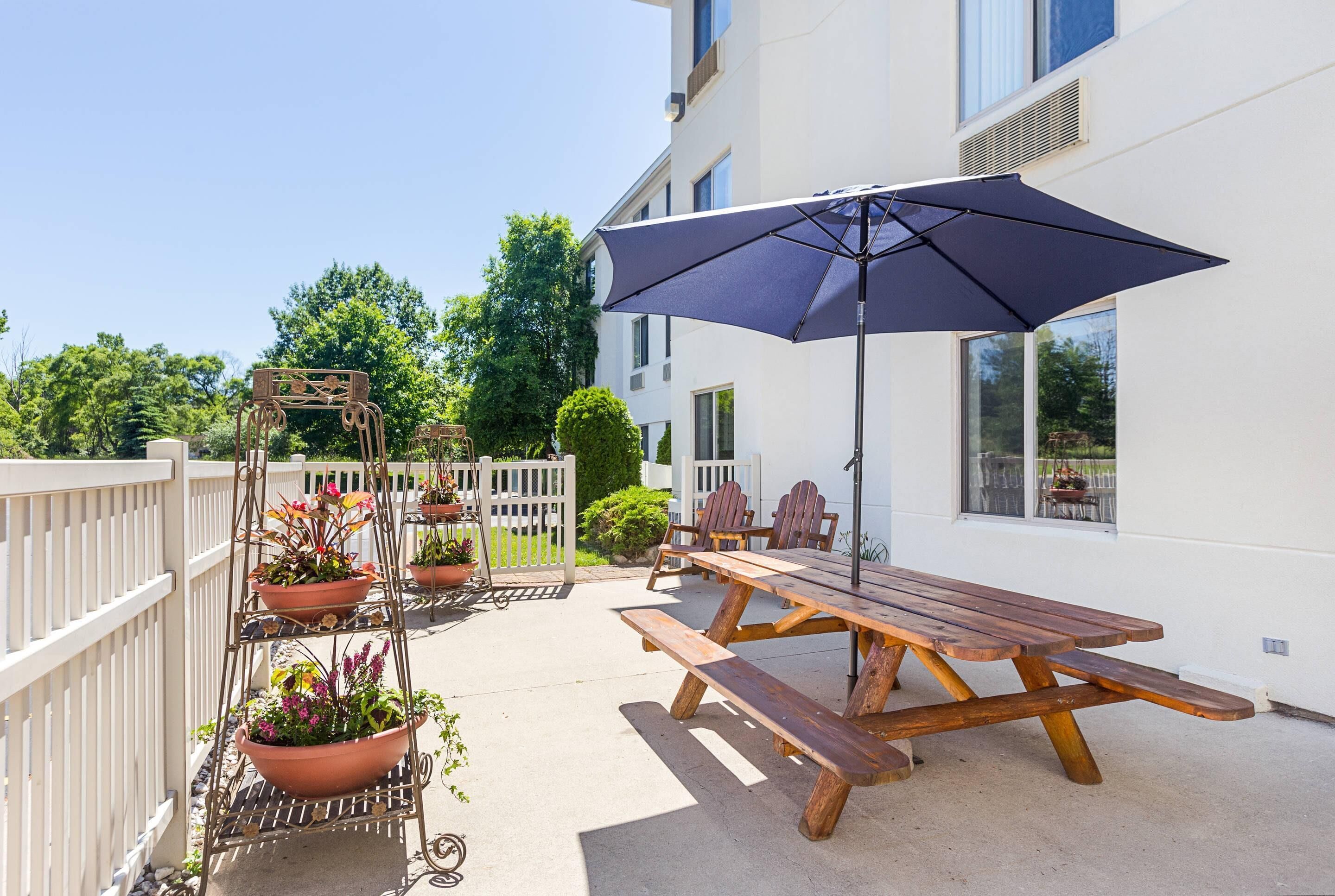 A wooden picnic table with an umbrella, potted plants, and a white fence.