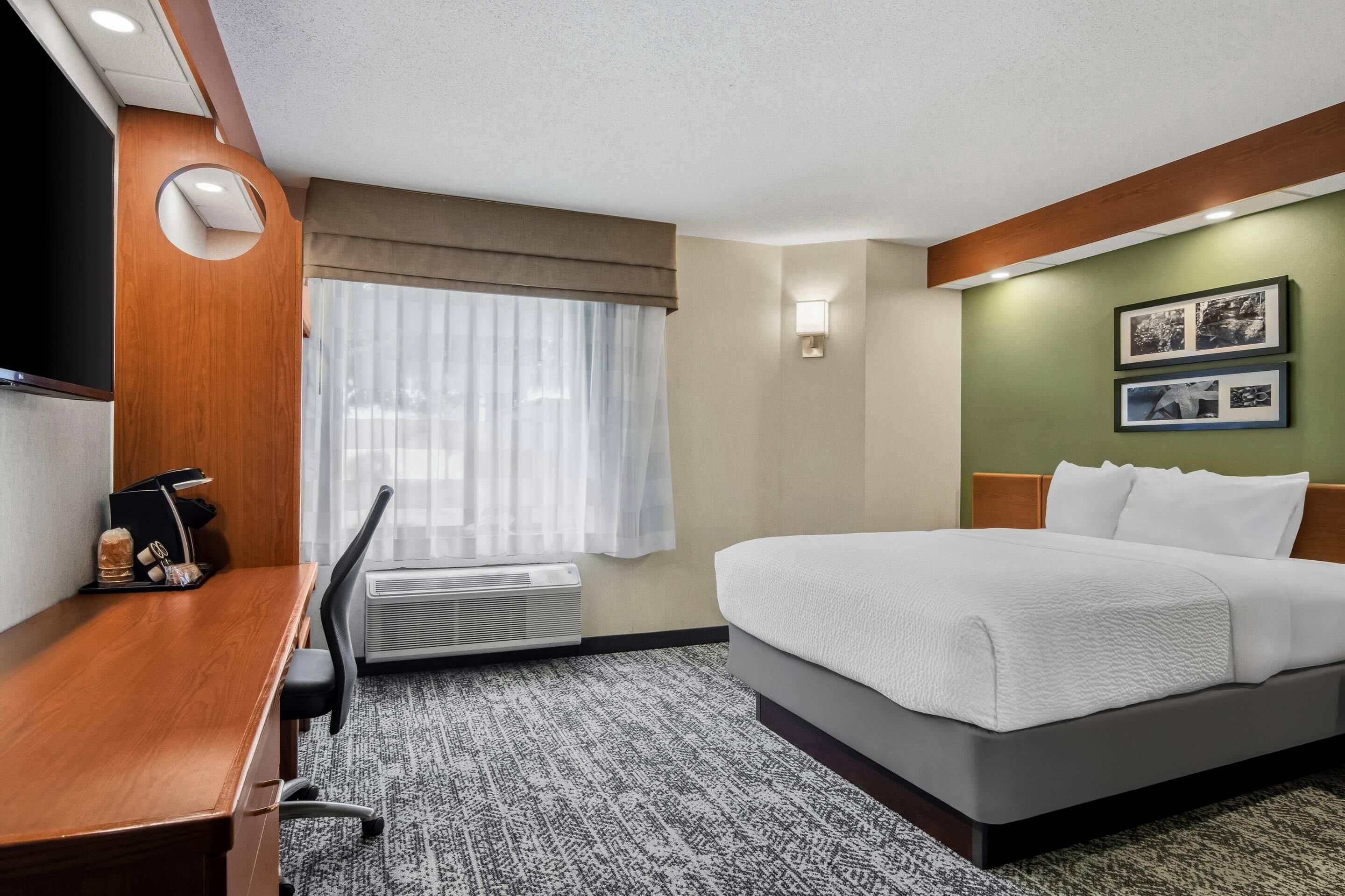 A hotel room with a bed, desk, chair, air conditioning unit, and a window with curtains.