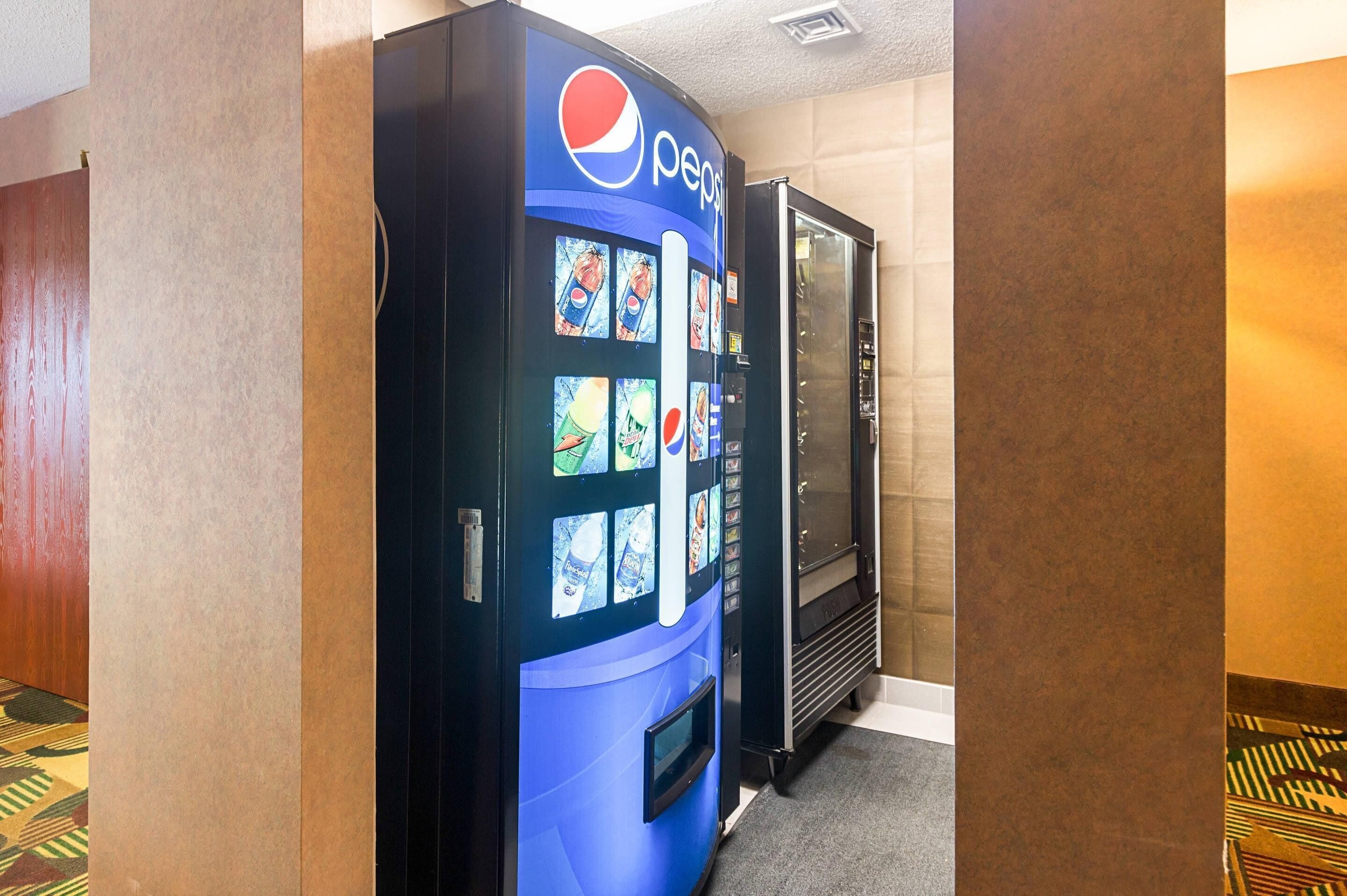A vending machine with a Pepsi logo, a refrigerator, and patterned carpet.