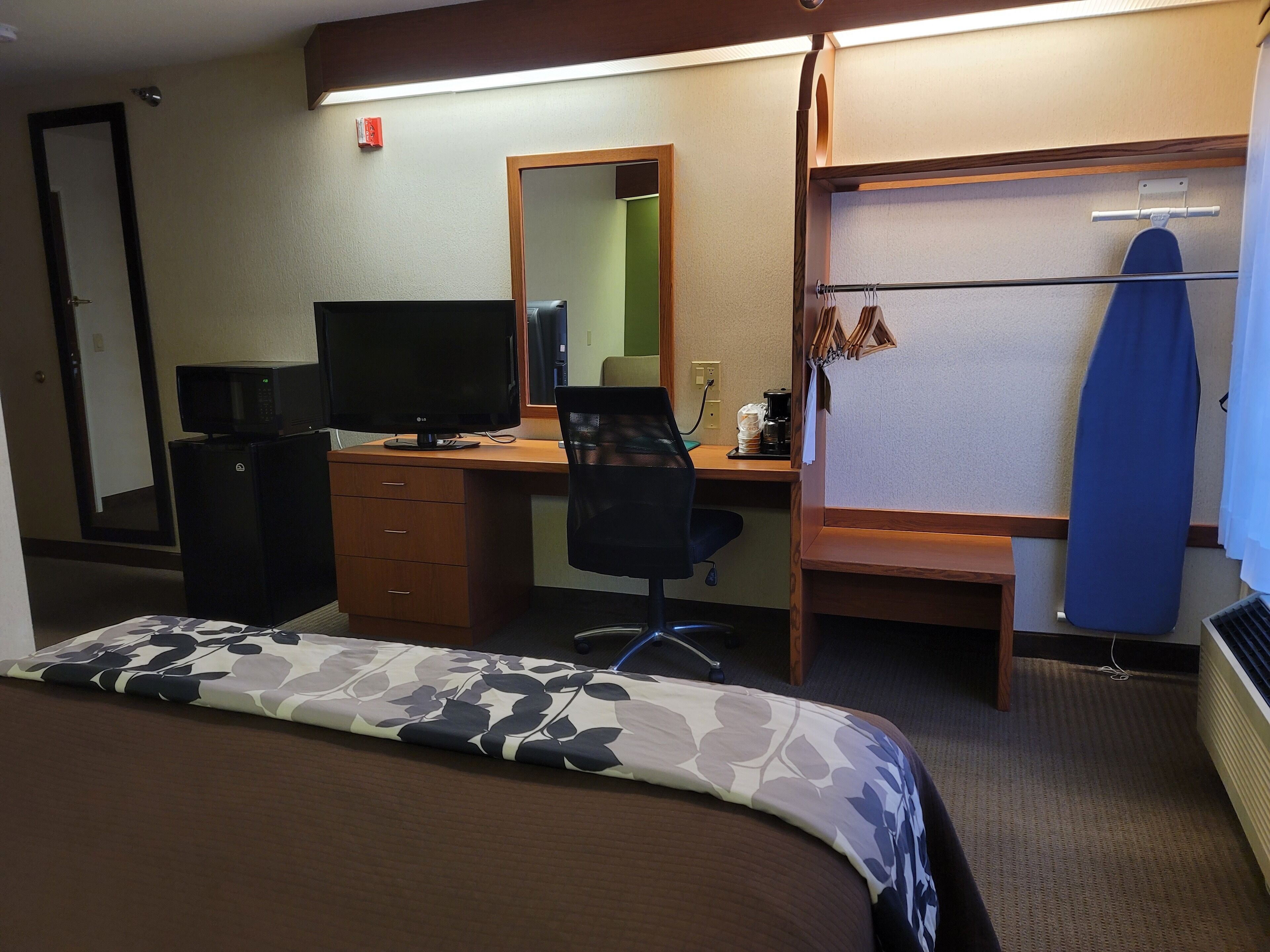 A hotel room with a bed, a desk with a computer, a chair, a mirror, a coat rack, and a heater.