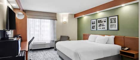 A hotel room with a bed, a desk with a computer, a chair, a TV, and a window with curtains.