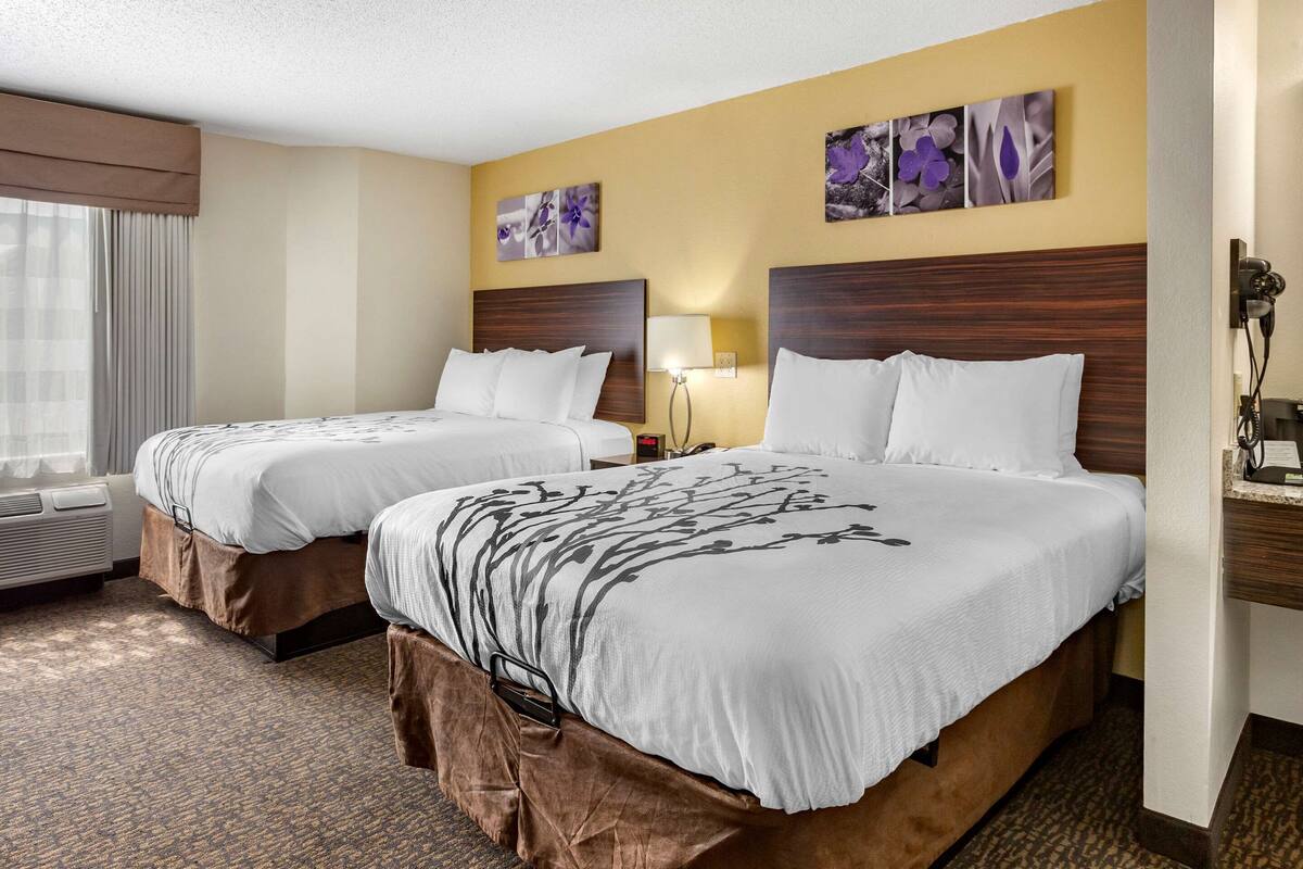 Quarto de hotel com duas camas, uma janela grande com cortinas, telefone fixado na parede e uma luminária.
