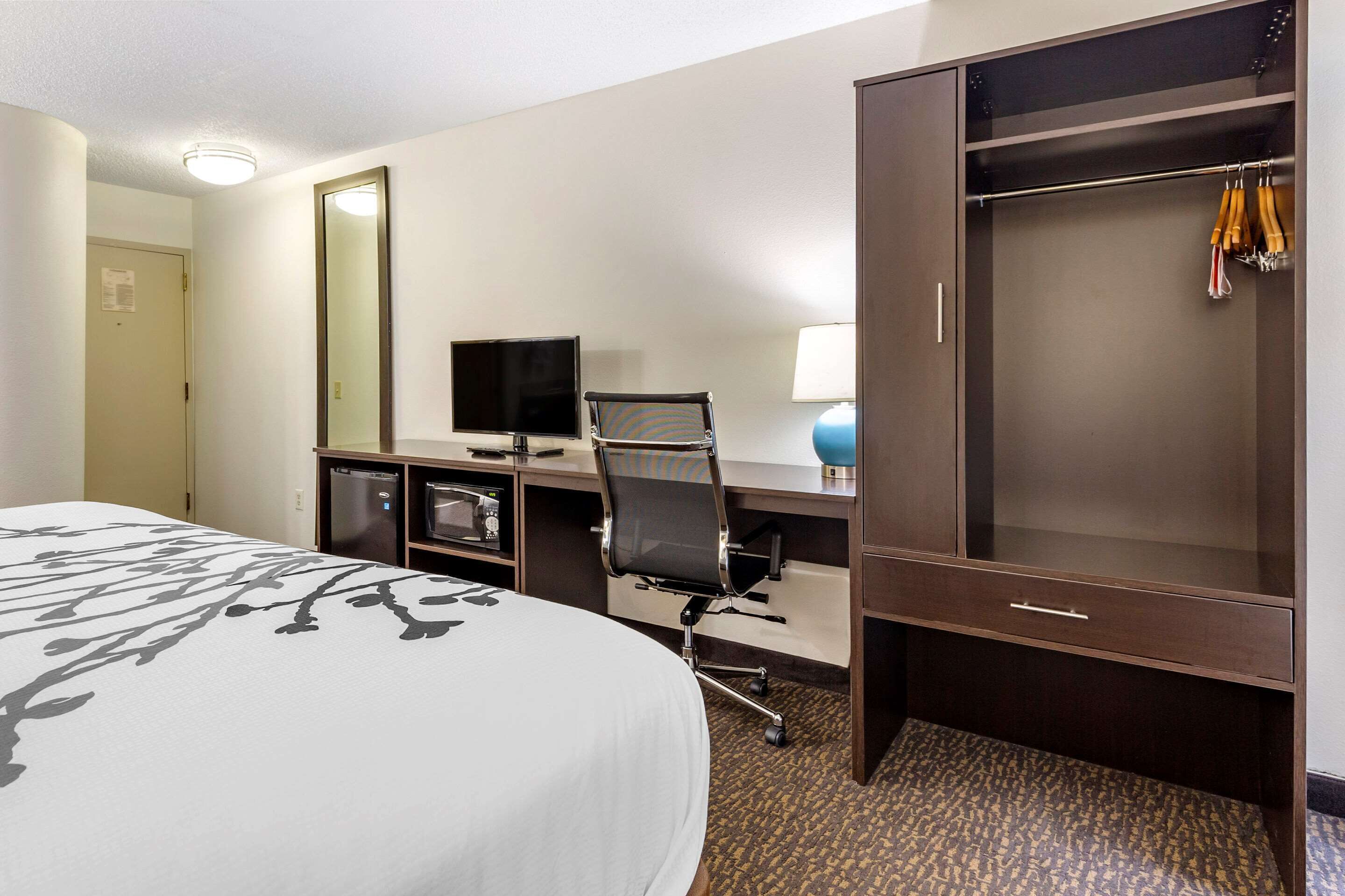 A hotel room with a bed, a desk with a computer, a chair, a wardrobe, and a mirror.