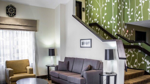 A living room with a sofa, armchair, and coffee table, a staircase with a green leaf pattern, and a clock on the wall. - Sleep Inn Garner - Clayton (Garner)
