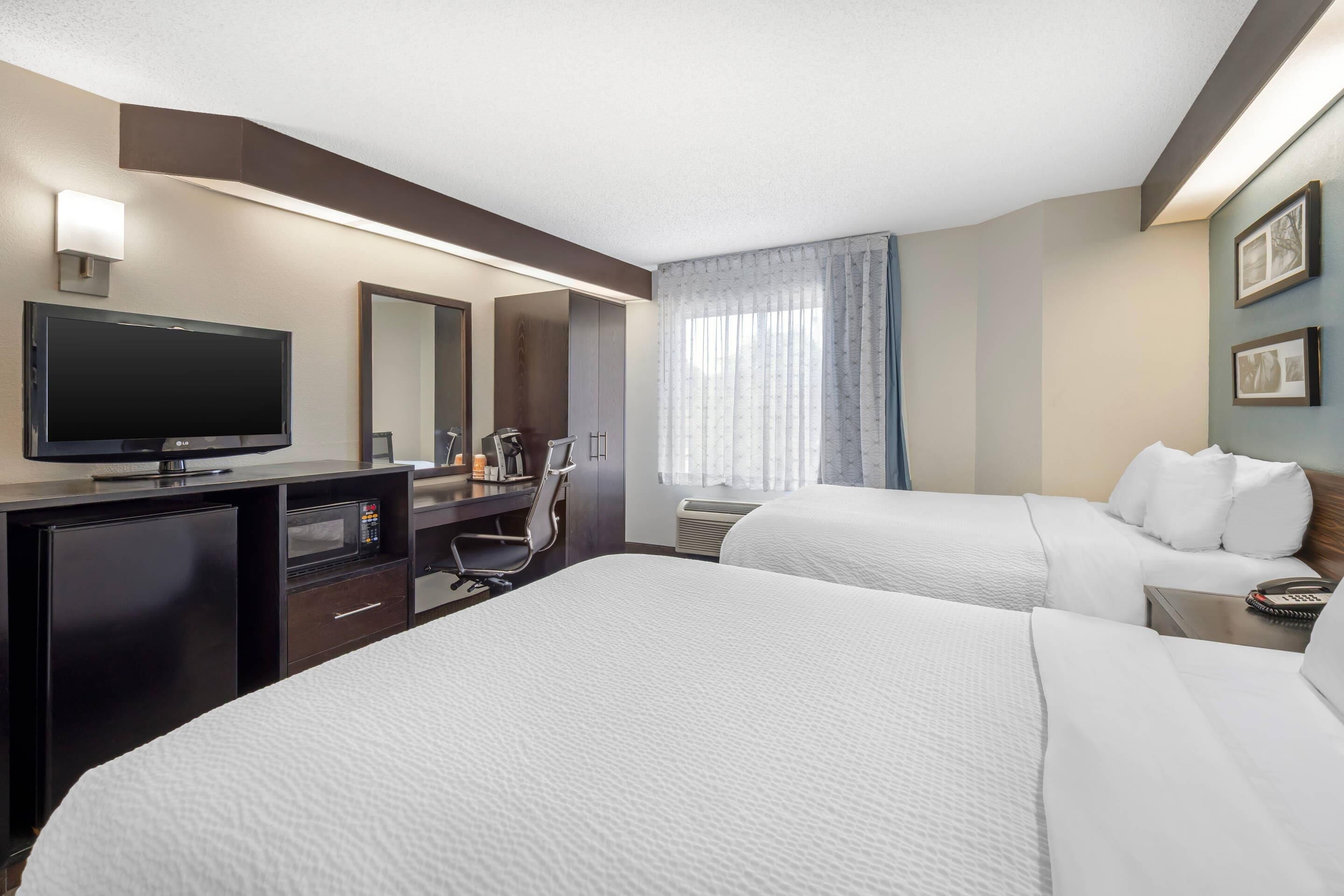 Ein Hotelzimmer mit zwei Betten, einem Schreibtisch mit Flachbildfernseher, einem Sessel, einer Schreibtischlampe, einem Heizkörper und einem Fenster mit Vorhängen.