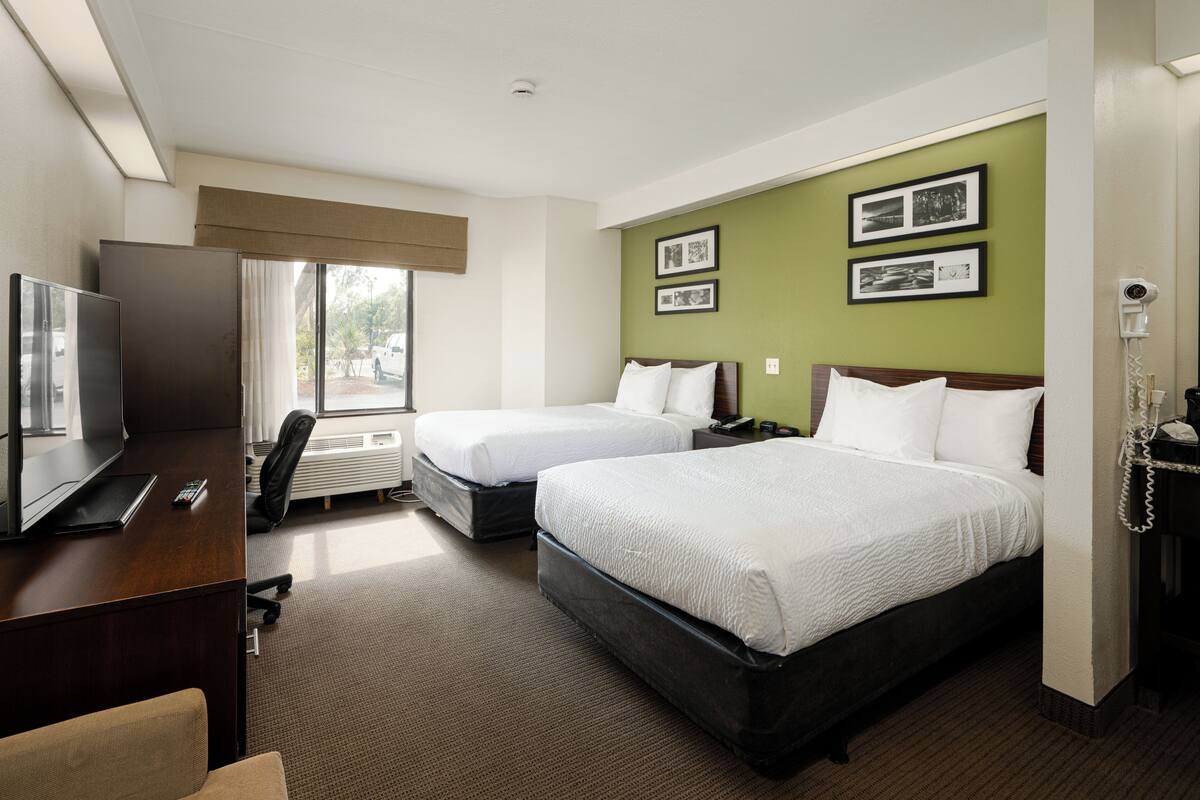 A hotel room with two beds, a desk, a TV, and a window with a view of the outside.