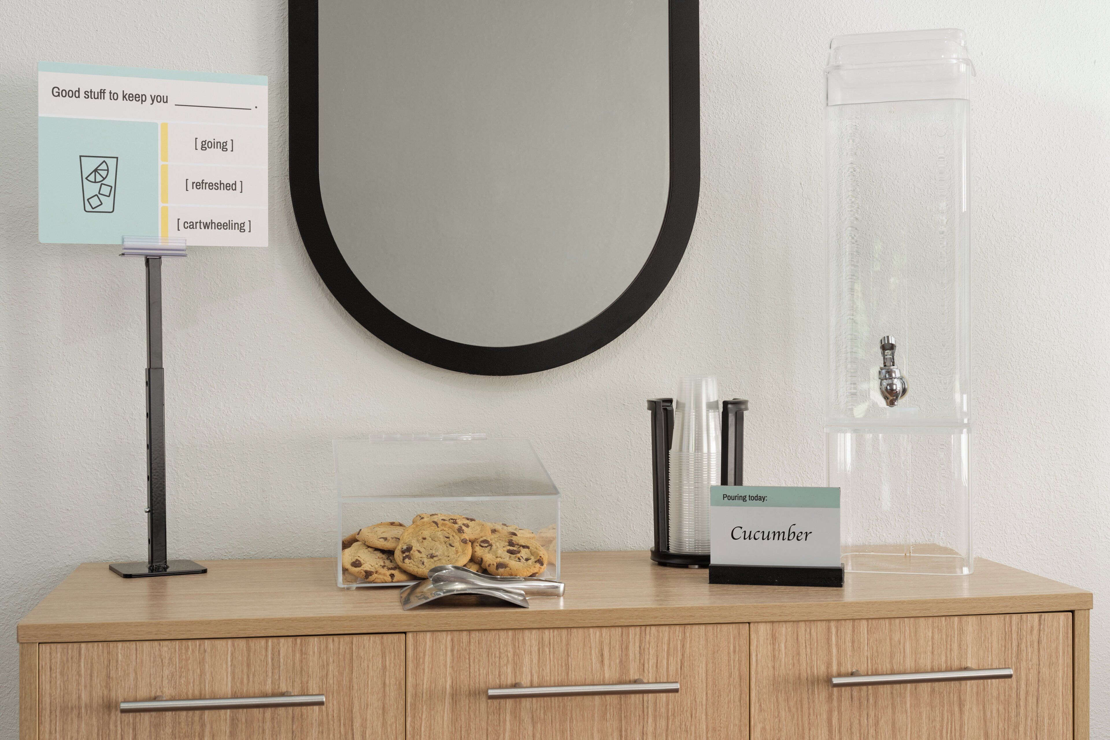 Una barra de madera con un letrero, un recipiente transparente de galletas, un dispensador de pepinos y un espejo con marco negro.