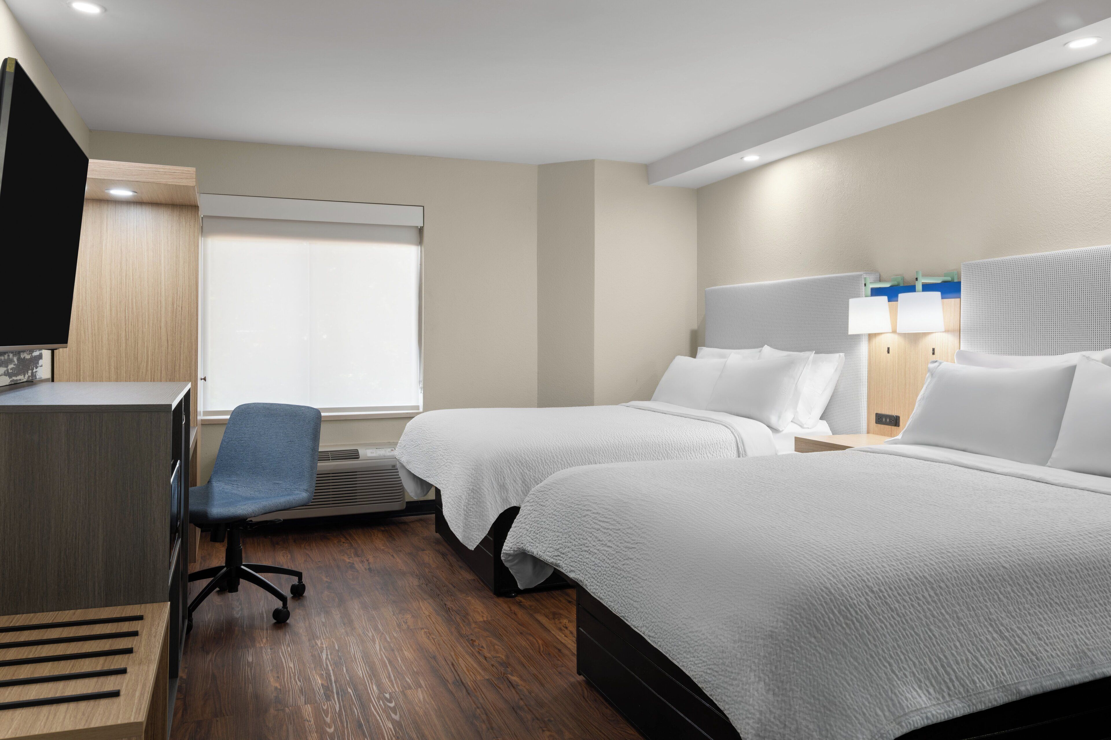 A hotel room with two beds, a desk, a chair, and a TV.
