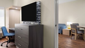 A modern hotel room with a flat-screen TV mounted on a cabinet, a desk with a chair, a small side table with a lamp, and a sofa. - Garner Hotel Aiken South by IHG (Aiken)