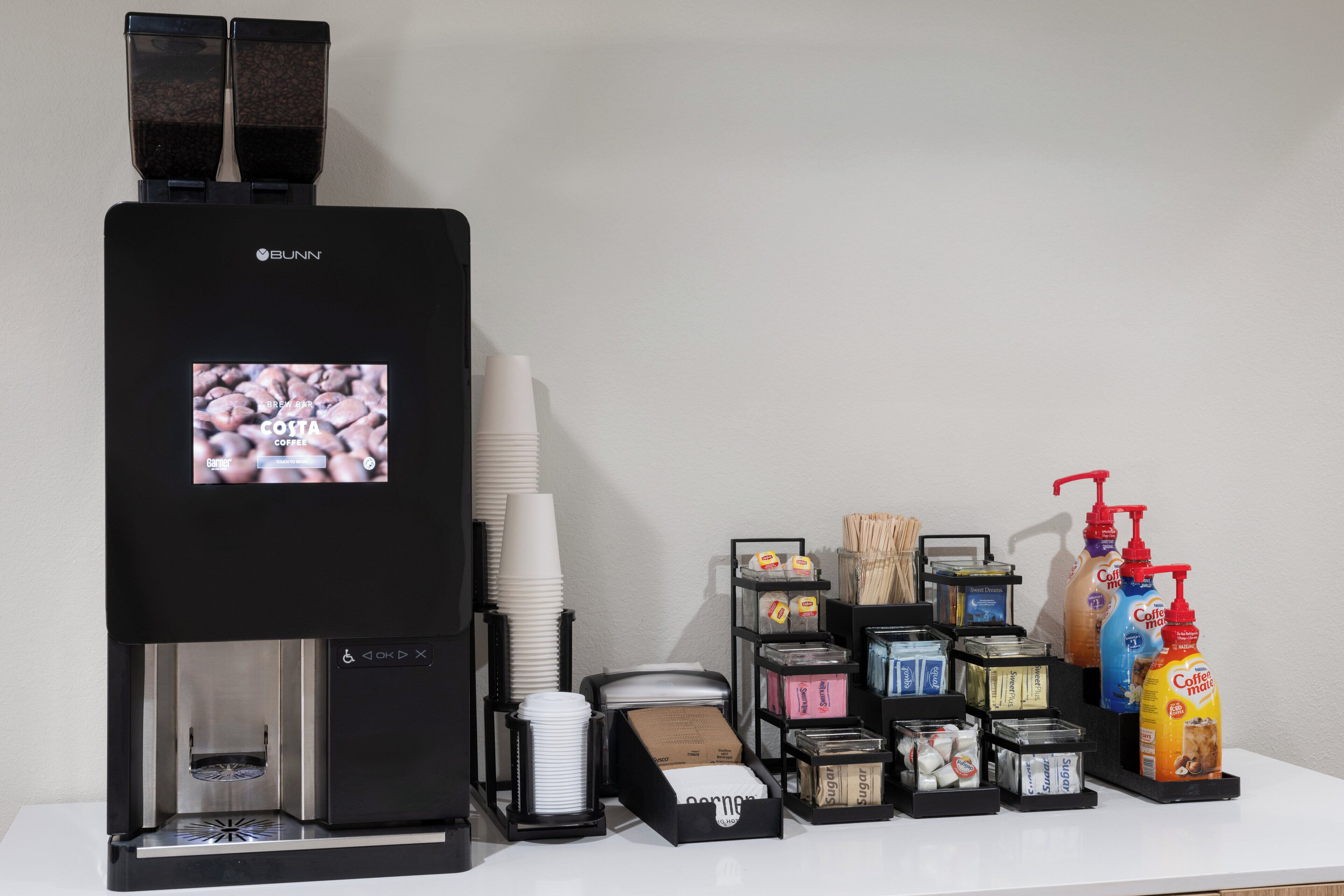 A coffee machine with a screen displaying coffee beans, surrounded by various coffee-related items like cups, sugar, and creamer.
