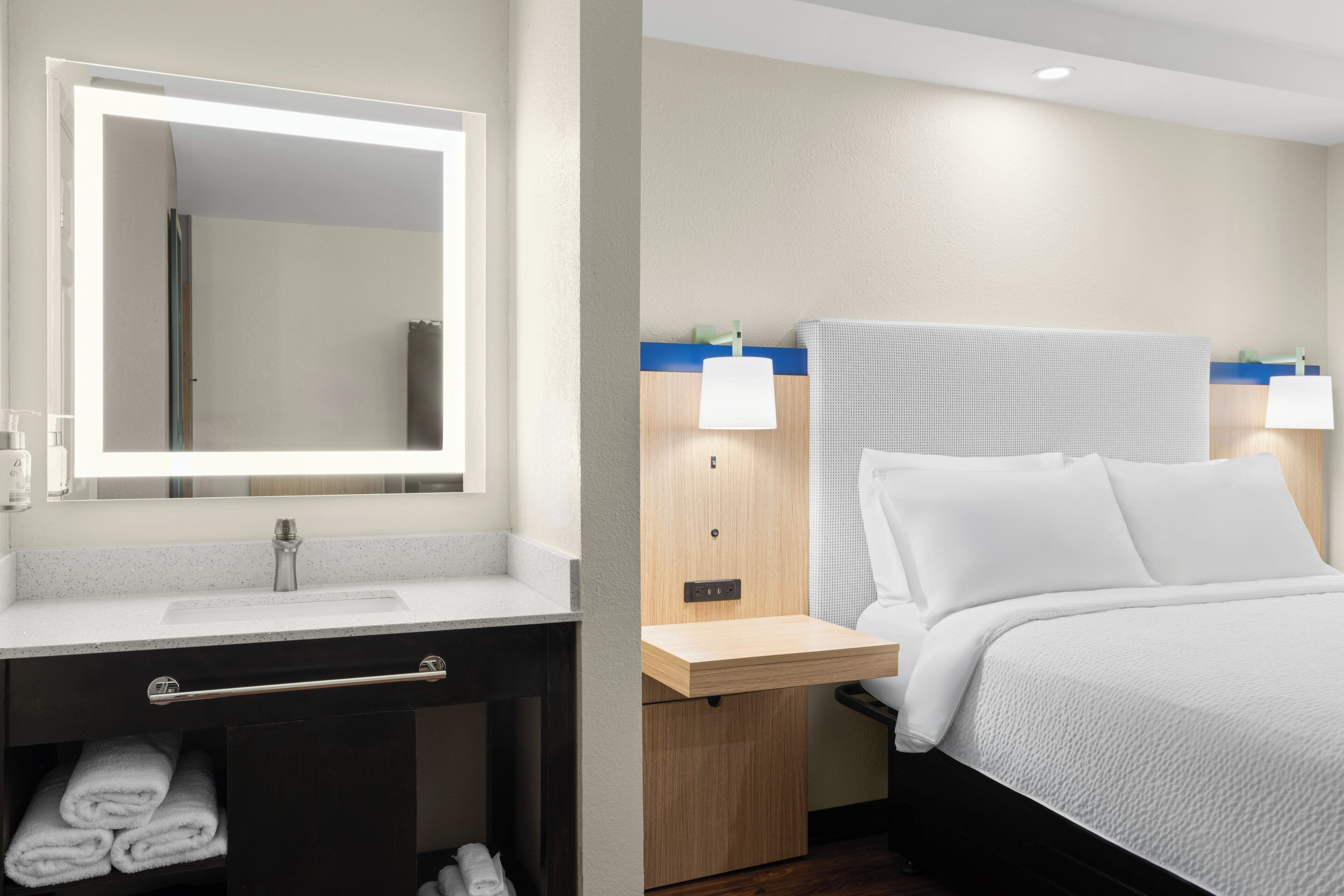 Ein Hotelzimmer mit Bett, Nachttisch mit Lampe, Spiegel, Waschbecken und Handtuchhalter.