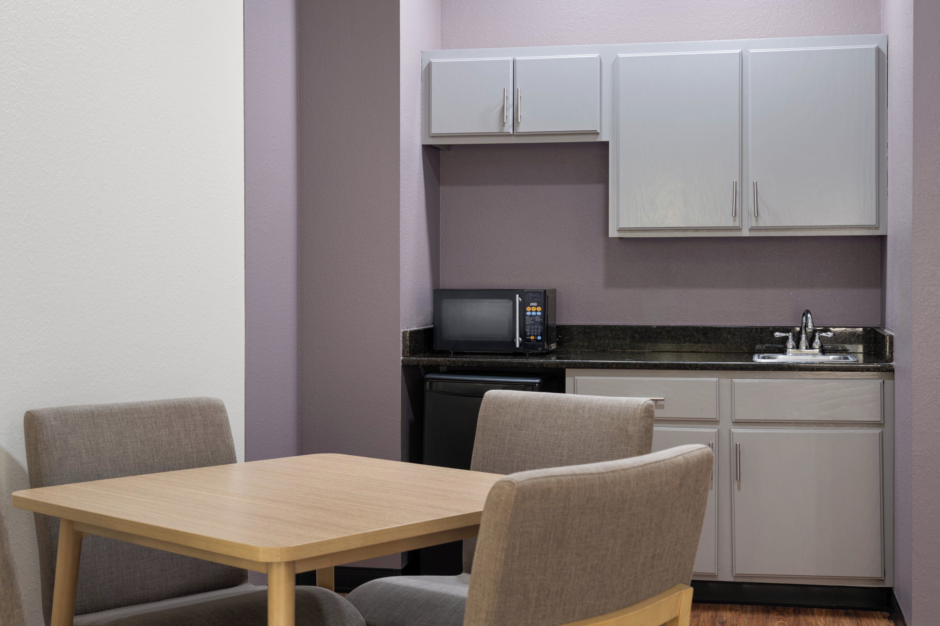 Een kleine kitchenette met een magnetron, spoelbak en kastjes. Er staat een tafel met twee stoelen.