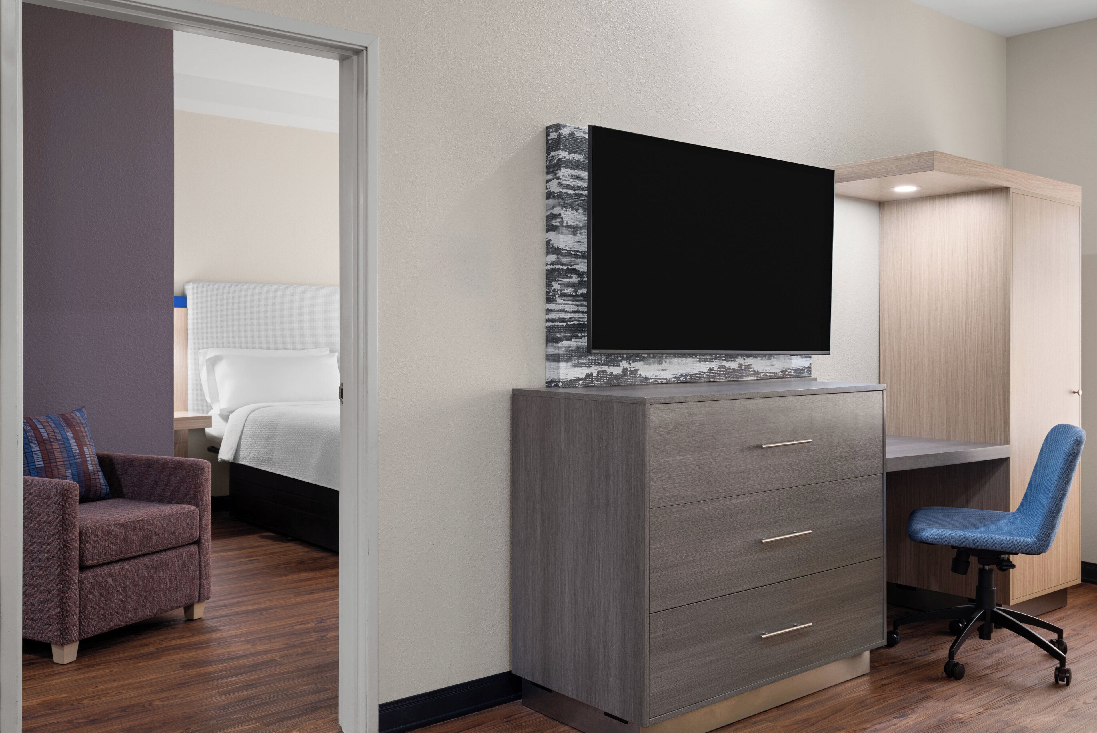 Ein modernes Hotelzimmer mit einem großen Flachbildfernseher an einem grauen Schrank, einem Holztisch mit Stuhl, einem Bett mit weißer Bettwäsche und einem kleinen Sessel.