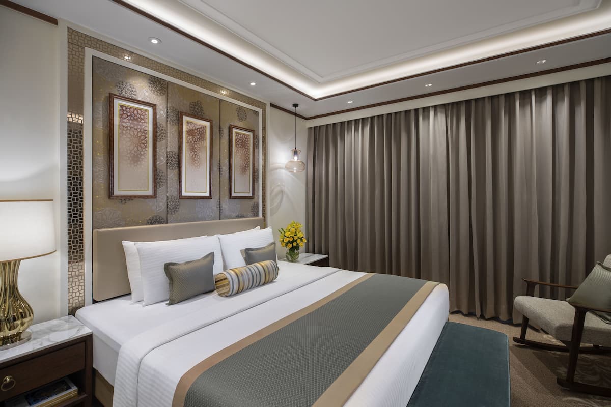 A modern hotel room with a large bed, a nightstand with a lamp, a chair, and a view of the city through curtains.