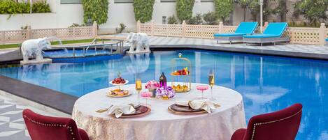 A poolside dining area with a table set for two, surrounded by a pool and lounge chairs.