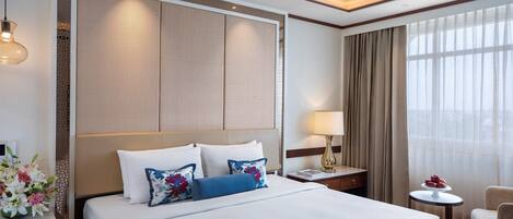 Ein Hotelzimmer mit einem großen Bett, einem Nachttisch, einer Lampe, einem Sessel und Blick ins Freie durch das Fenster.