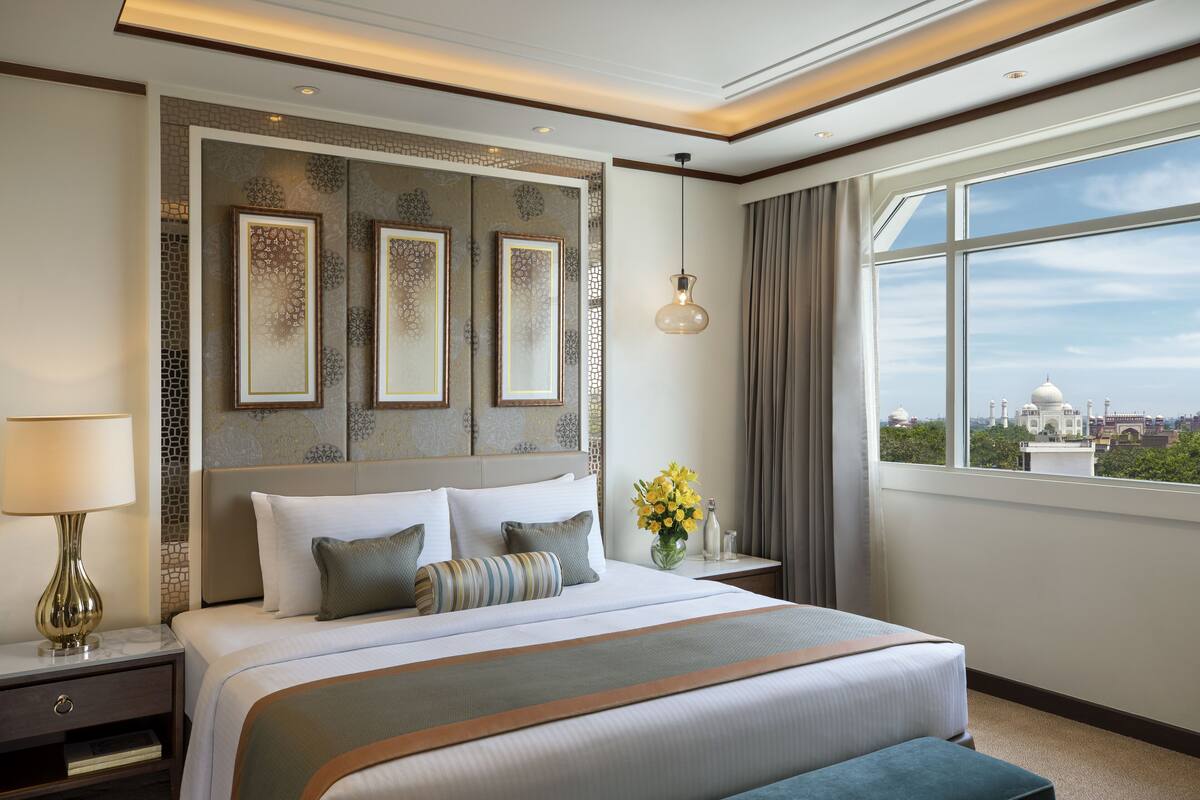 A hotel room with a large bed, a nightstand with a lamp, a vase of flowers, and a view of a white building through the window.