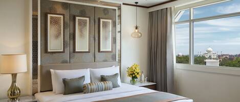 Habitación de hotel con una cama grande, una mesita de noche con una lámpara, un jarrón de flores y vistas a un edificio blanco a través de la ventana.