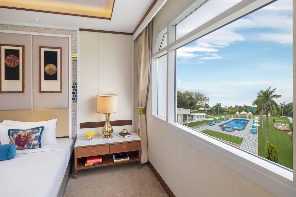 A hotel room with a bed, bedside table, lamp, framed artwork, and a view of a swimming pool and garden.
