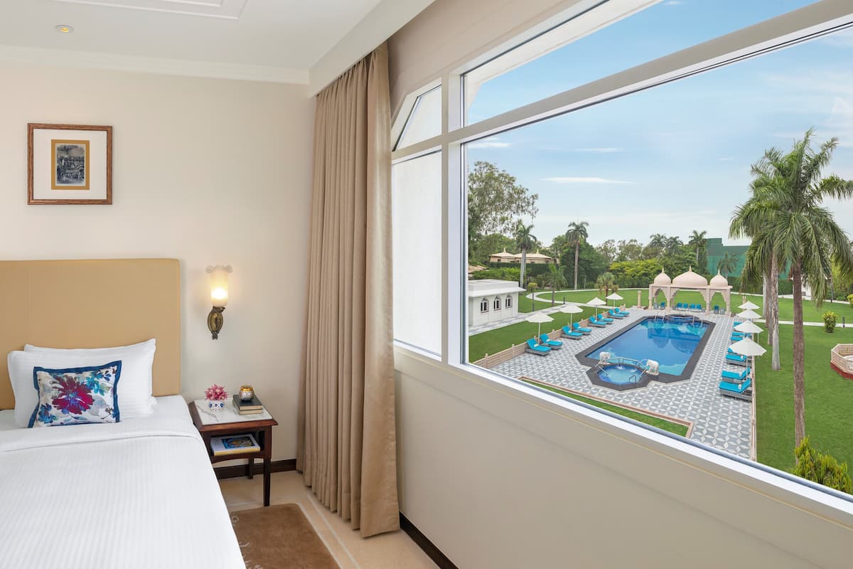 A hotel room with a large window, a bed, a nightstand, and a view of a pool and garden.