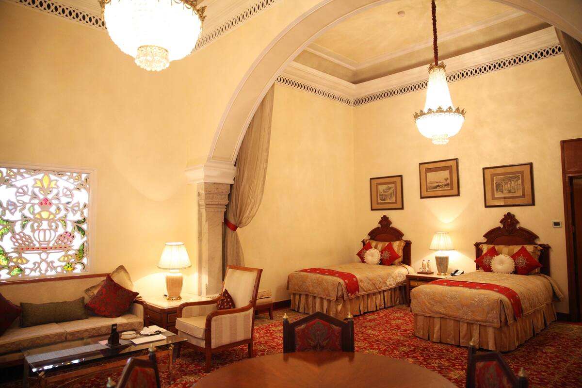 A hotel room with two beds, a sofa, a dining table, and a chandelier.