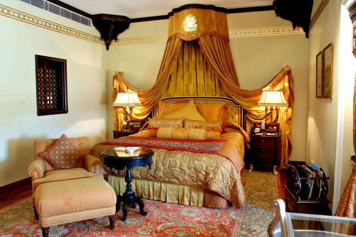 A traditional bedroom with a four-poster bed, a sitting area with a chair and ottoman, and a nightstand with a lamp.