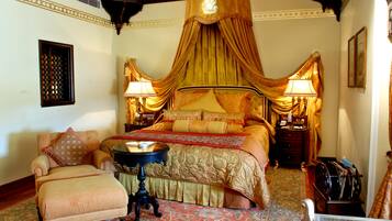 A traditional bedroom with a four-poster bed, a sitting area with a chair and ottoman, and a nightstand with a lamp.