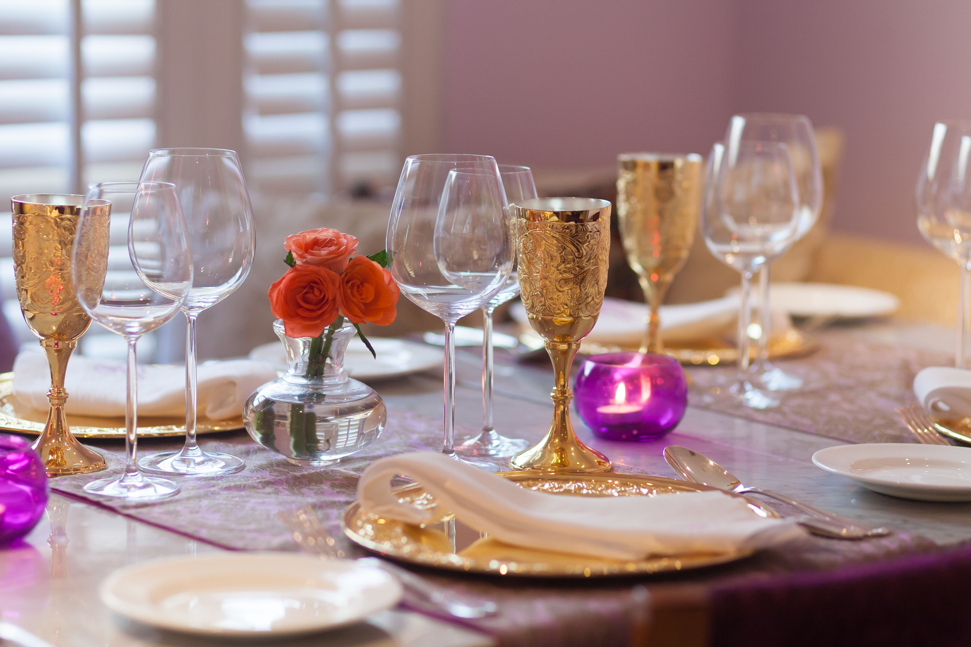 Uma mesa posta com taças de vidro transparente, um cálice dourado e um pequeno vaso com uma única rosa.