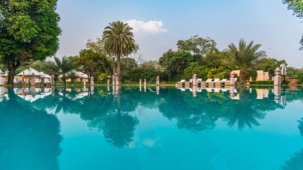 Une piscine aux eaux cristallines, bordée de chaises longues et d’ombrelles, entourée d’arbres et d’un bâtiment au style architectural traditionnel.
