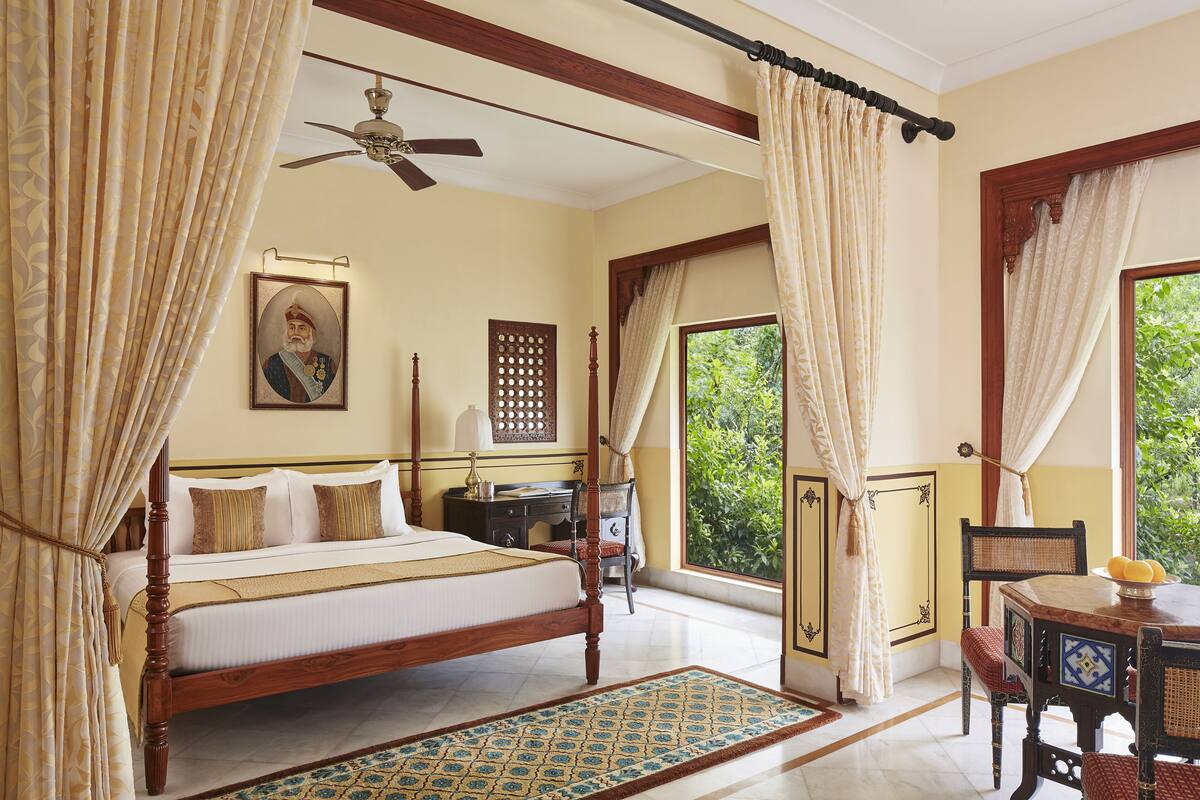A bedroom with a four-poster bed, a ceiling fan, a desk with a chair, and a window with curtains.
