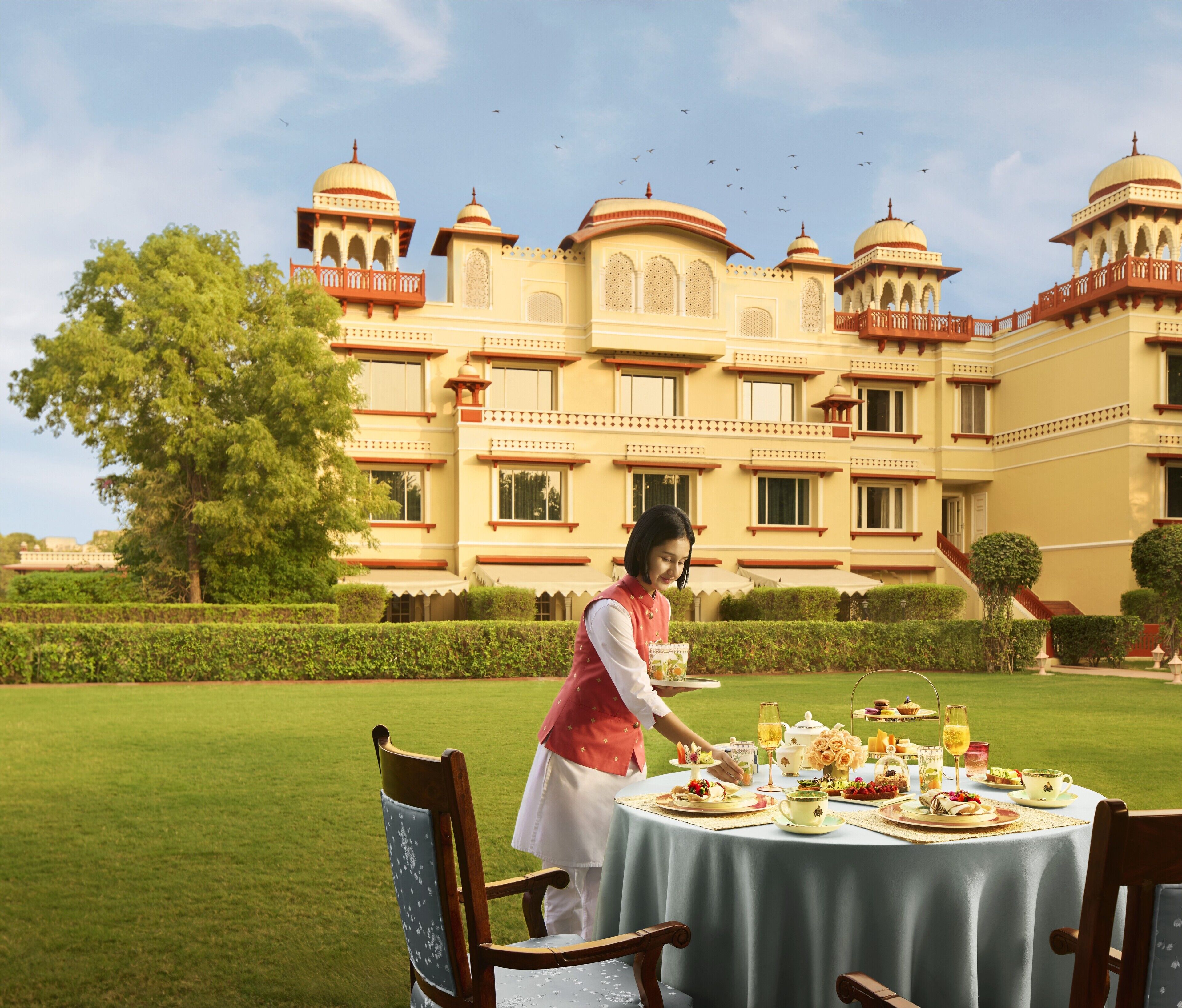 A dining area with a table set for a meal, a person serving, and a large building with domes in the background.