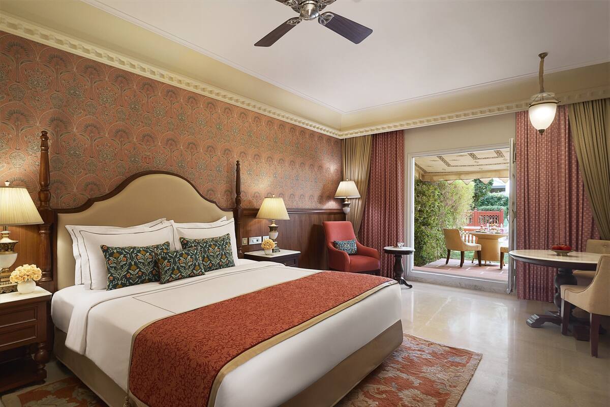 A hotel room with a large bed, two bedside lamps, a red chair, a small table, and a view of the outdoors.