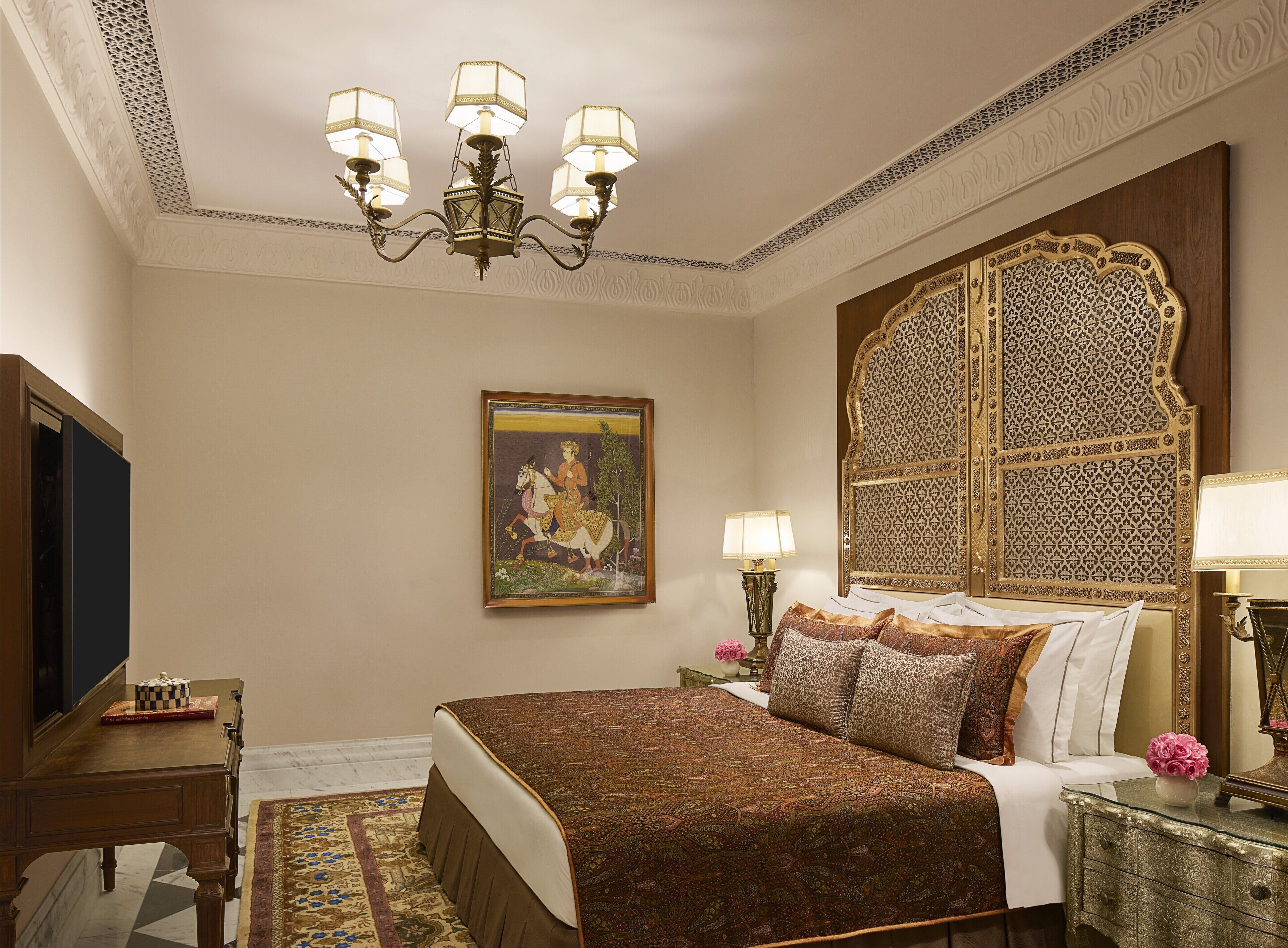 Quarto com uma cama grande, cabeceira elaborada, um lustre, um quadro emoldurado e uma mesa de cabeceira com abajur.