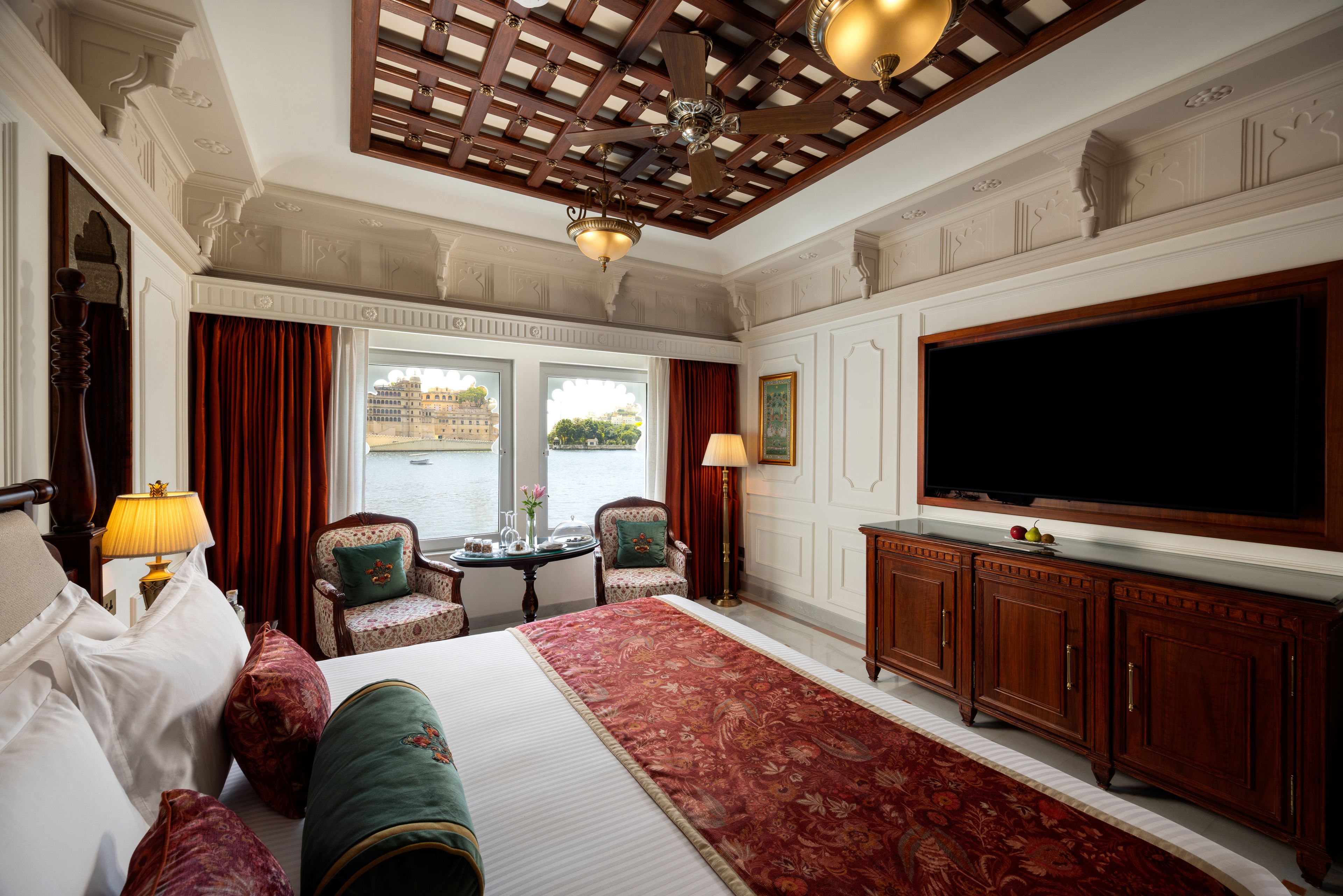 A hotel room with a large bed, a television, a desk with a lamp, and a view of a body of water through the windows.