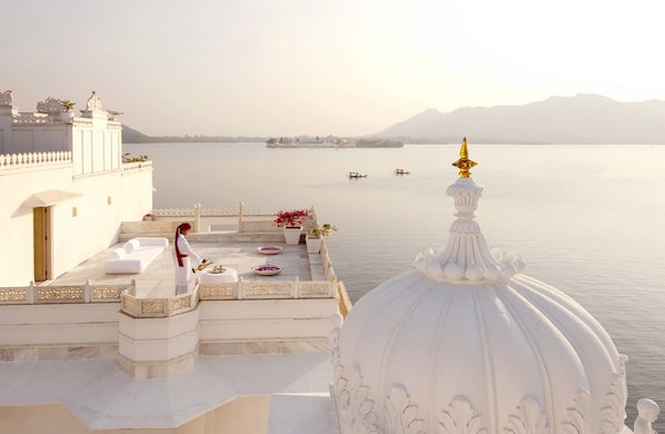 Una terraza con vistas a un lago tranquilo y montañas lejanas, que presenta un edificio blanco con detalles ornamentales y una cúpula blanca.