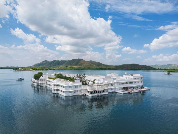 A large, multi-story white building with multiple balconies, situated on a lake with boats and mountains in the background. - Taj Lake Palace (Udaipur)
