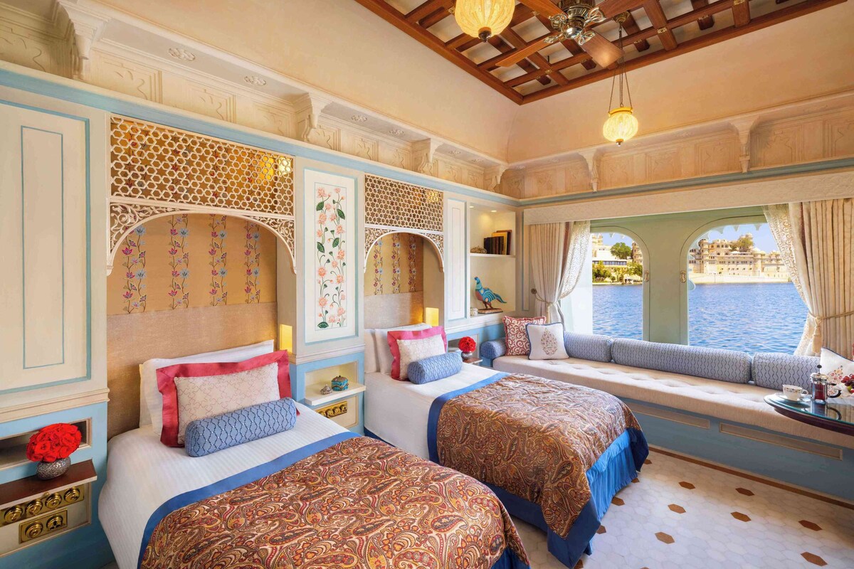 A hotel room with two beds, a window seat with a view of the water, decorative wall panels, and a chandelier.