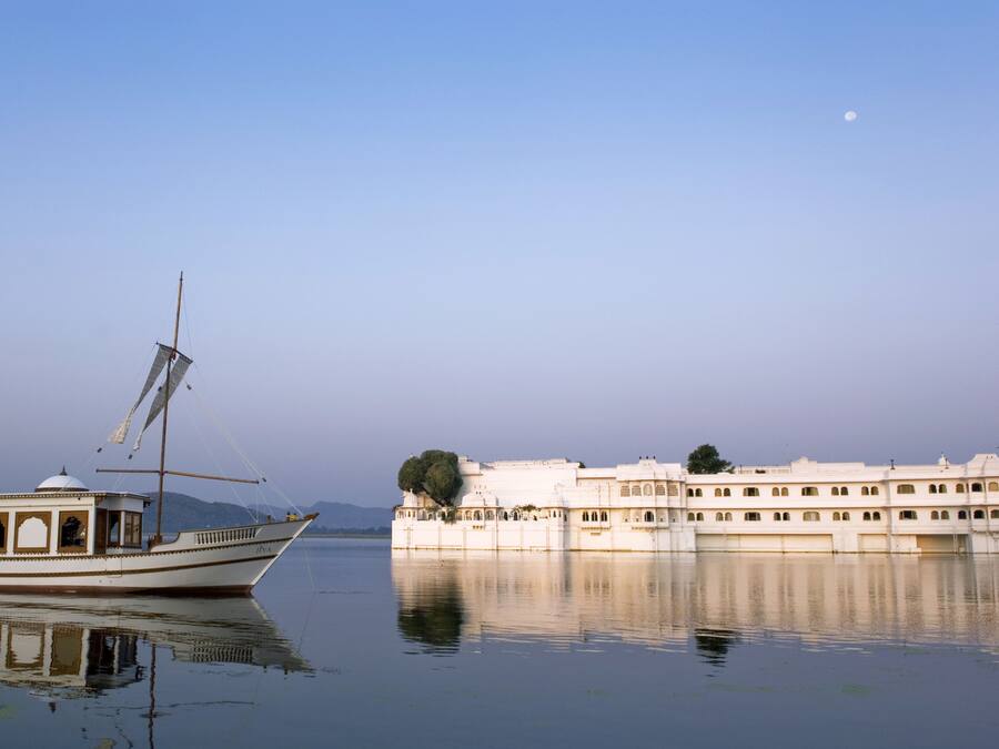 A boat with a mast and sails on calm water, a large white building with arched windows, and a clear sky with a moon.