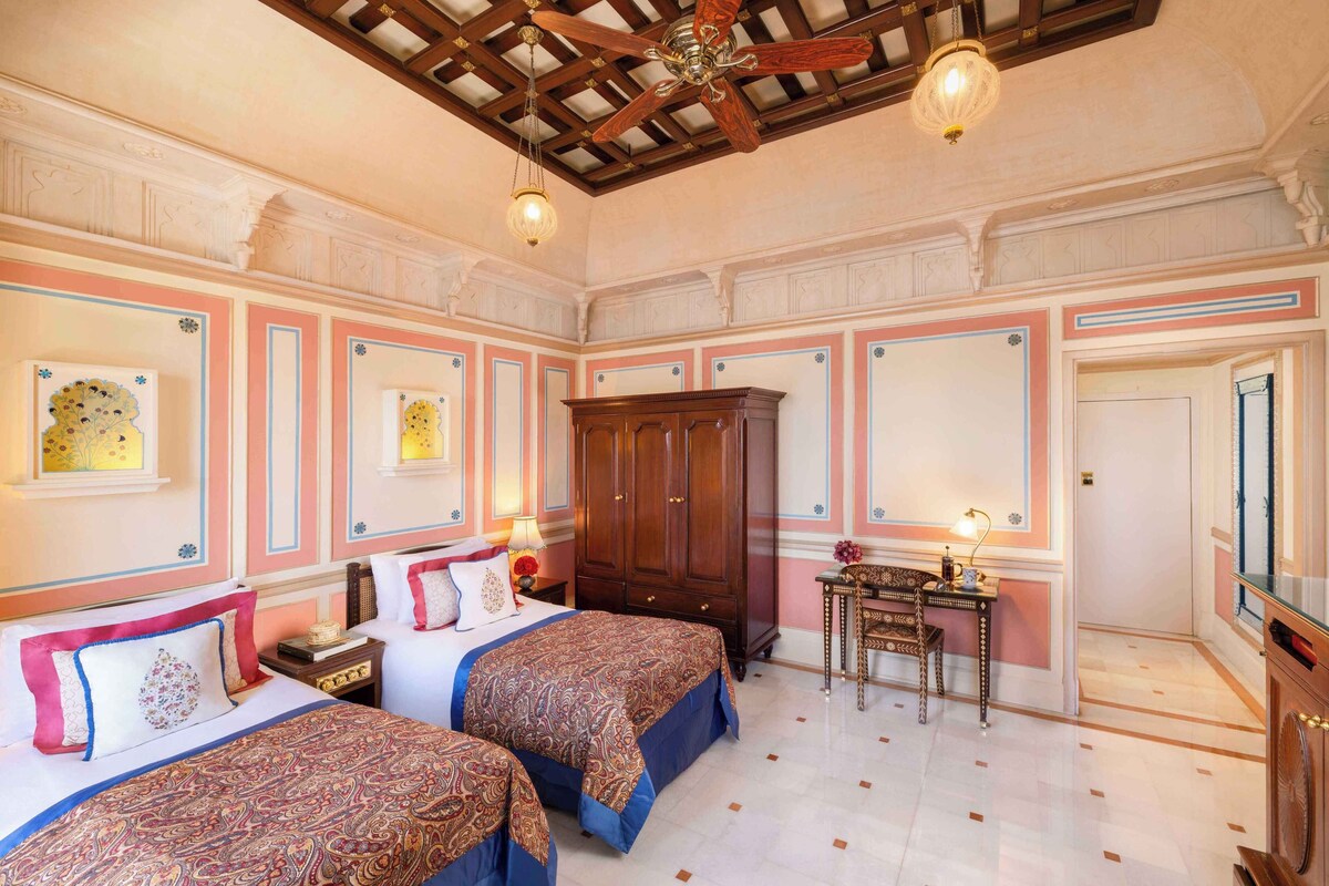 A hotel room with two beds, a wooden wardrobe, a desk with a chair, and a ceiling fan.