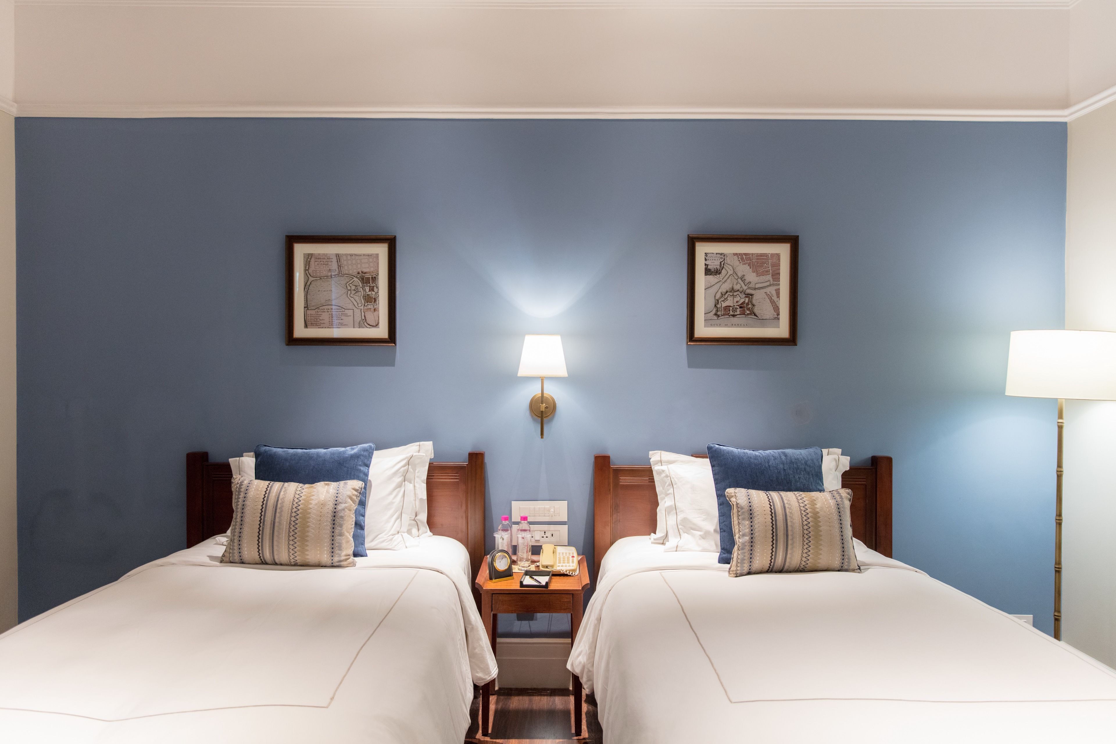 Habitación de hotel con dos camas, una mesita de noche, una lámpara y cuadros en las paredes.
