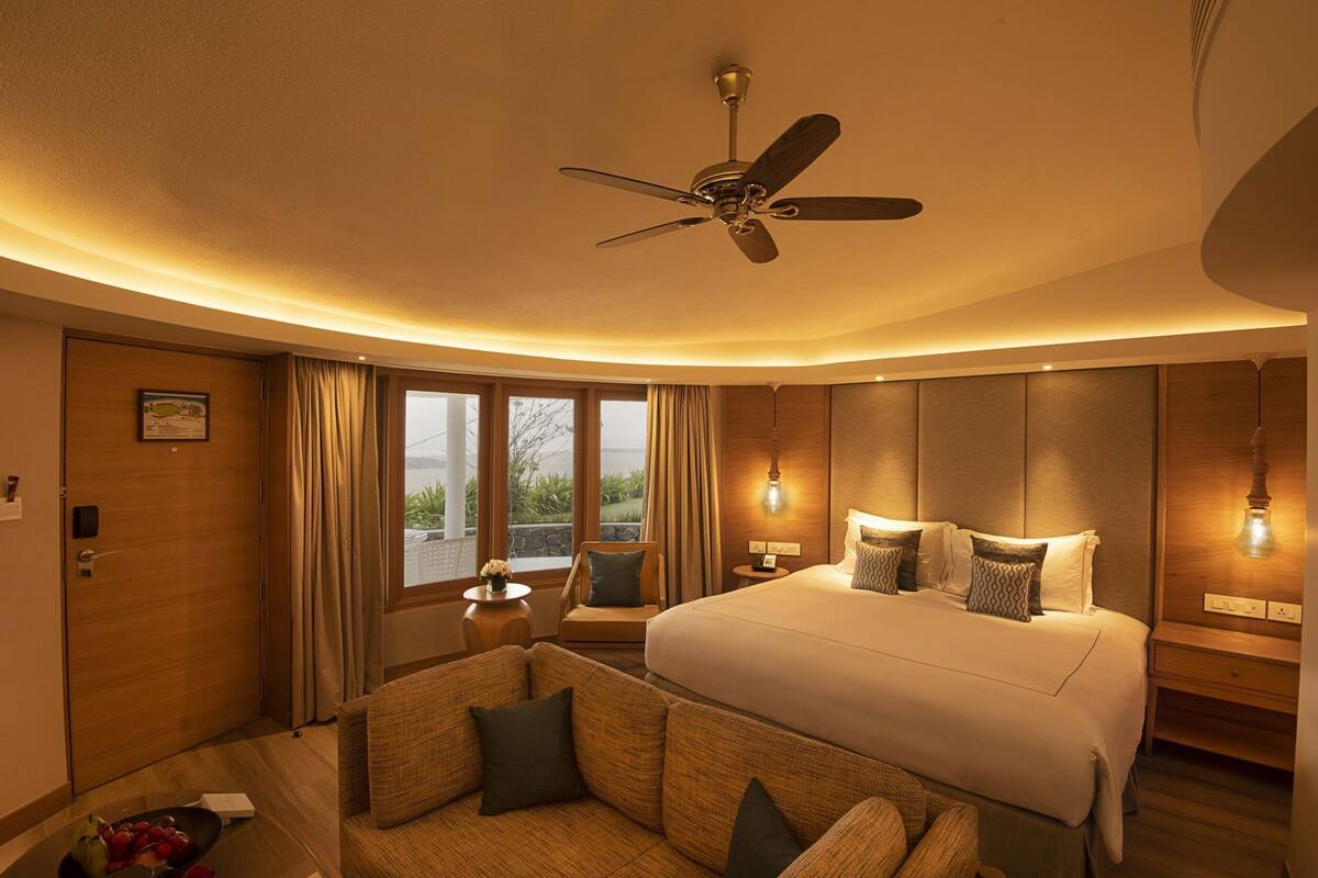 Habitación de hotel con cama, ventilador de techo, zona de estar con una silla, una mesita y vistas al exterior.