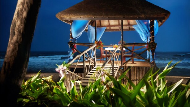 Een houten prieel met rieten dak, blauwe gordijnen en een tafel met stoelen aan het strand.