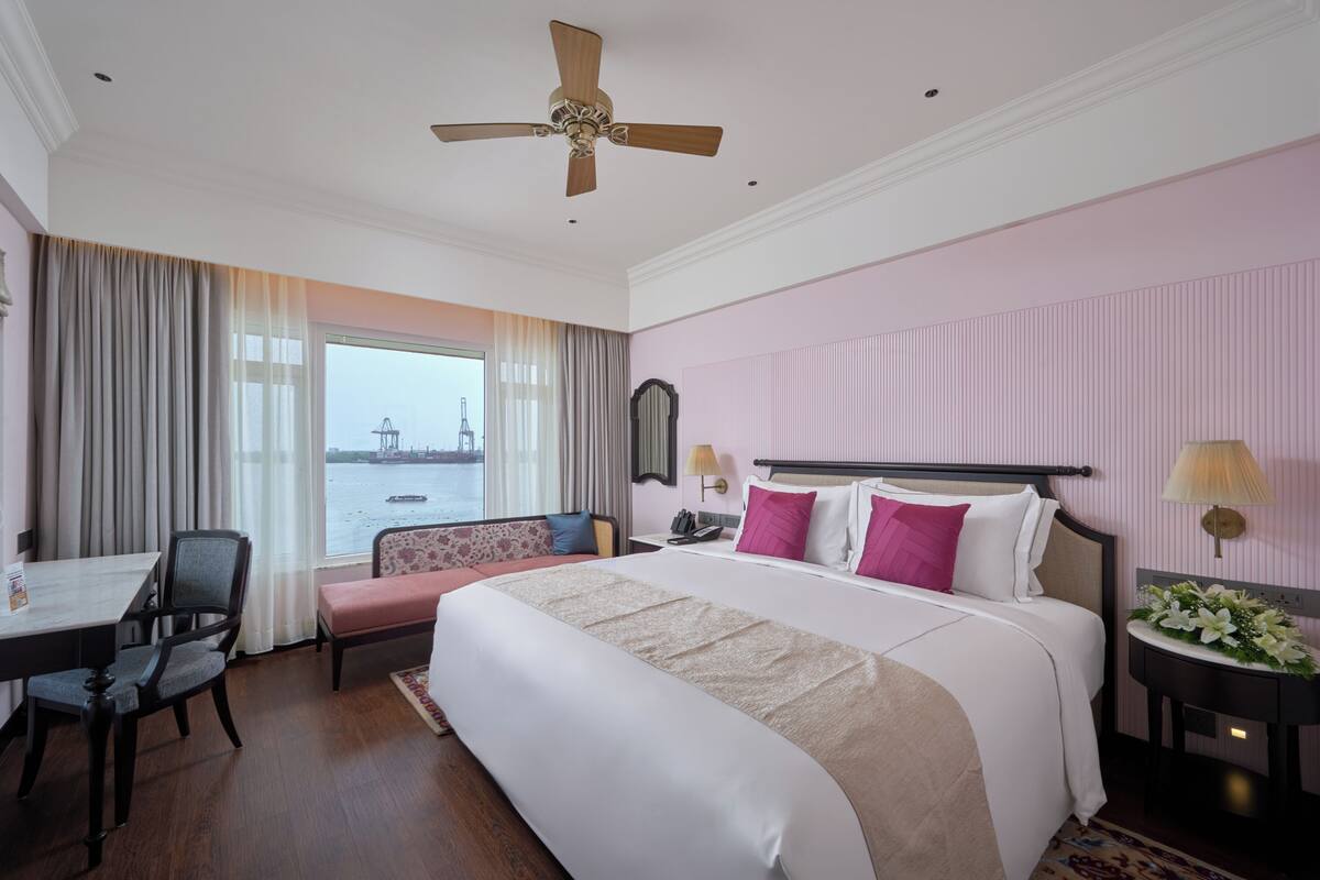 A hotel room with a bed, a desk, a chair, a window with a view of a body of water, and a ceiling fan.