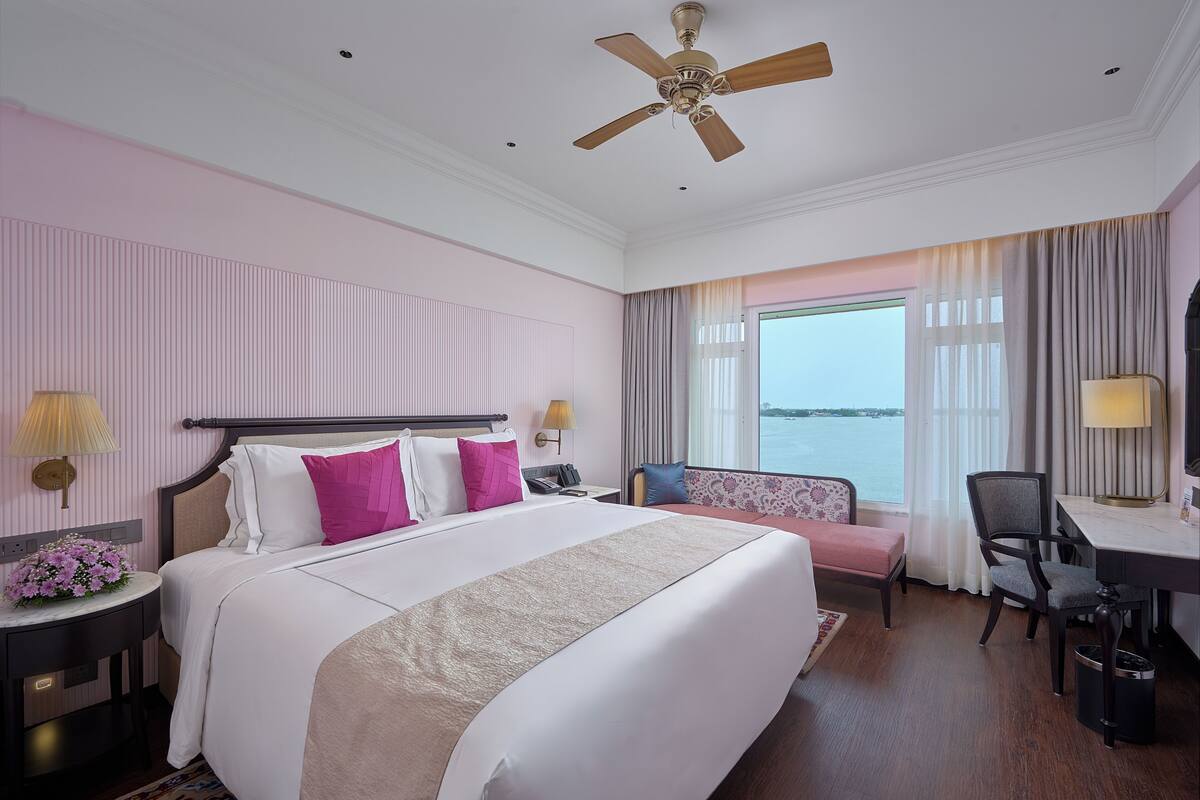 A hotel room with a large bed, a desk, a chair, a sofa, a ceiling fan, and a view of the sea.