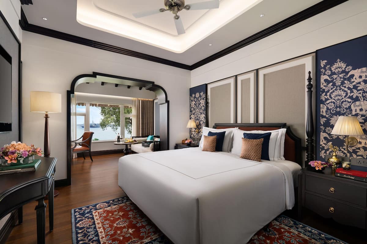 A spacious bedroom with a large bed, a desk, and a chair. There is a window with a view of a building and a chair outside.